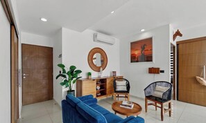 Uma sala de estar moderna com um sofá azul, duas poltronas, um armário de madeira, um espelho redondo e um quadro na parede.
