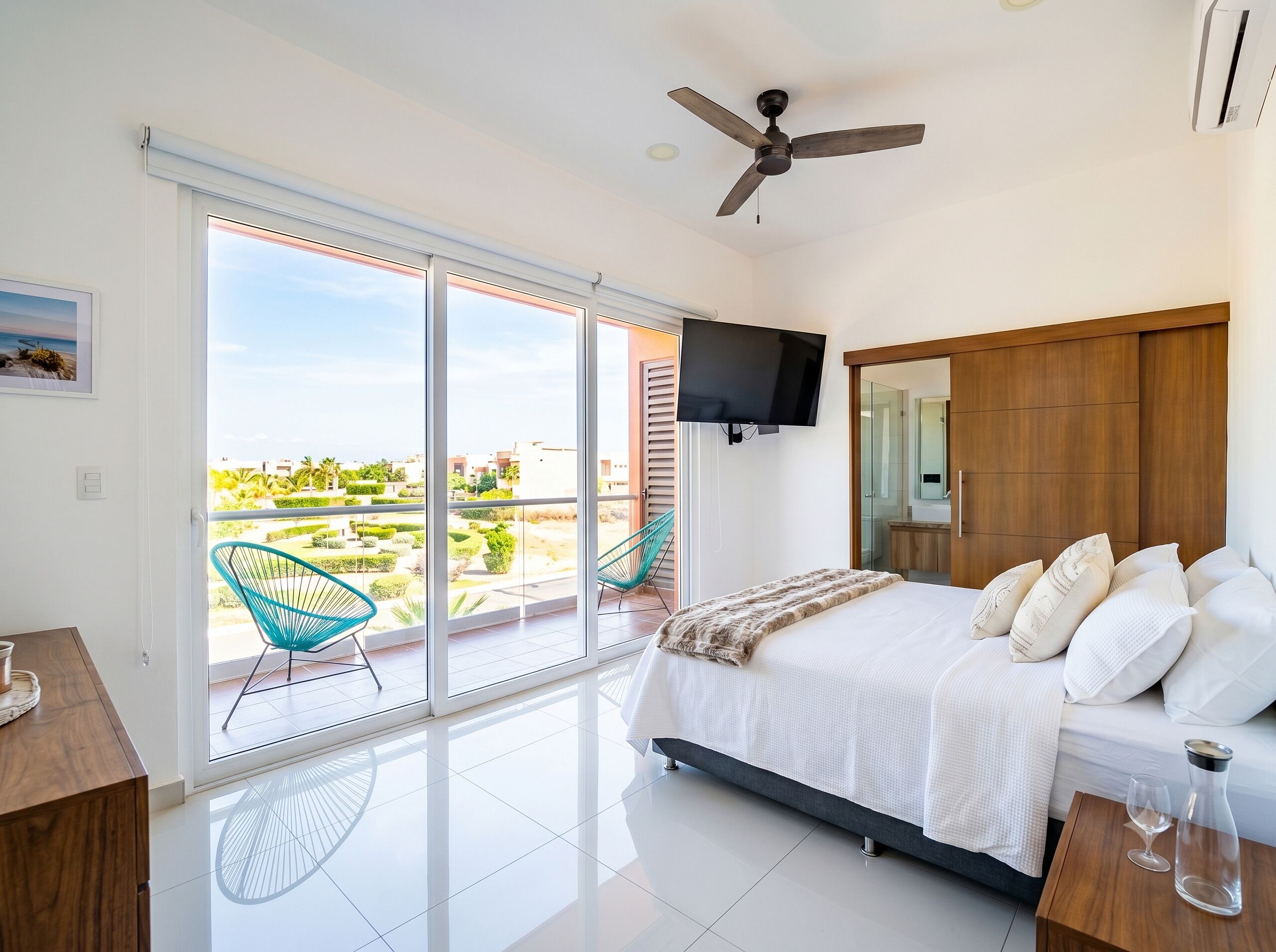 A bedroom with a bed, a TV, a ceiling fan, and a balcony with chairs.
