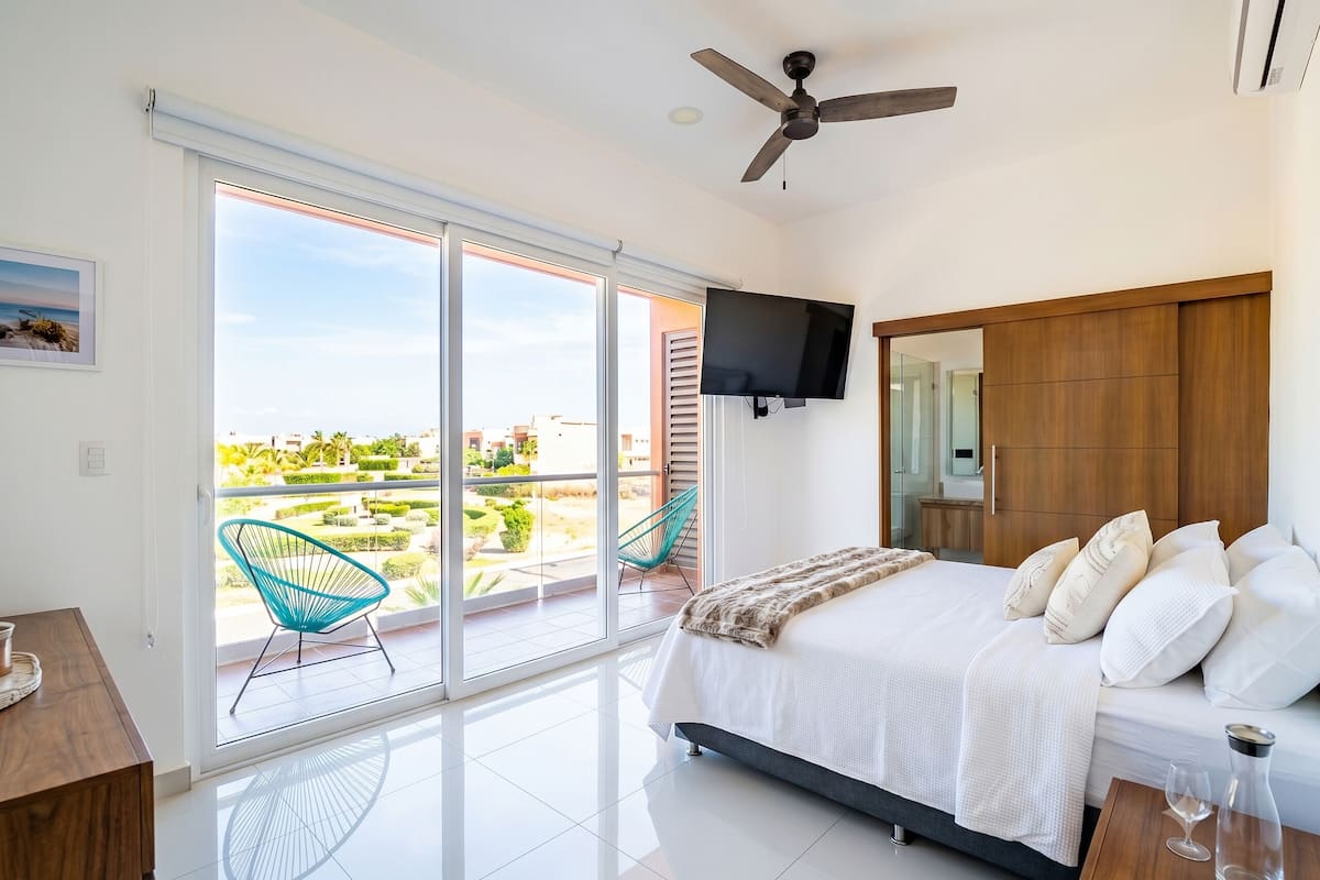 Un dormitorio con cama, televisión, ventilador de techo y balcón con sillas.