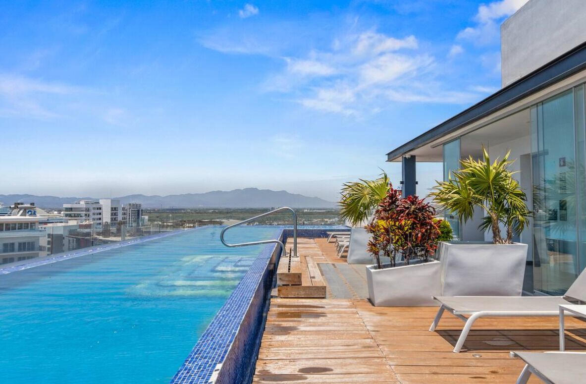 A rooftop infinity pool with a clear view of the ocean and distant mountains, surrounded by lounge chairs and potted plants.