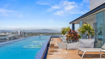 A rooftop infinity pool with a clear view of the ocean and distant mountains, surrounded by lounge chairs and potted plants.