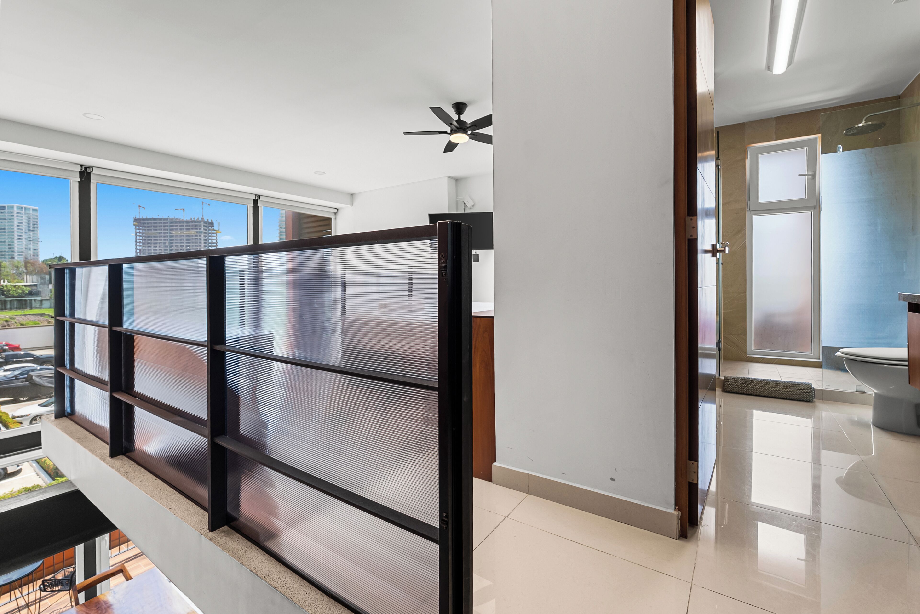 A modern interior with a view of the city through large windows, a ceiling fan, and a sliding glass door leading to a bathroom.