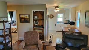 A living room with a patterned armchair, a black leather sofa, a wooden coffee table, and a kitchen in the background.
