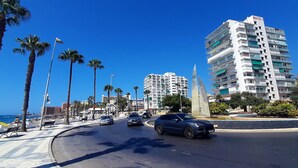 Una calle urbana costera con palmeras, un edificio moderno identificado como "MAITE 1", y un coche azul en movimiento.
