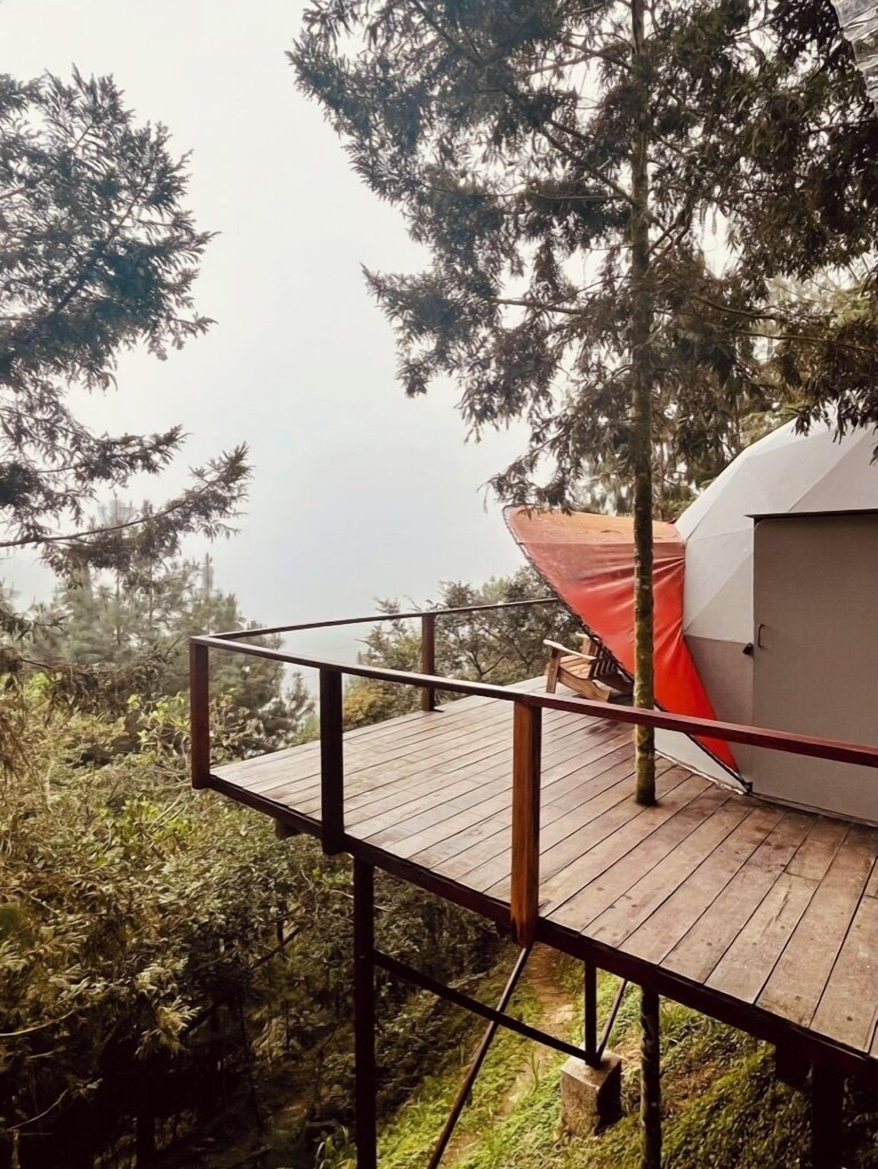 Una terraza de madera con una sombrilla roja, una carpa blanca y un bosque denso al fondo.