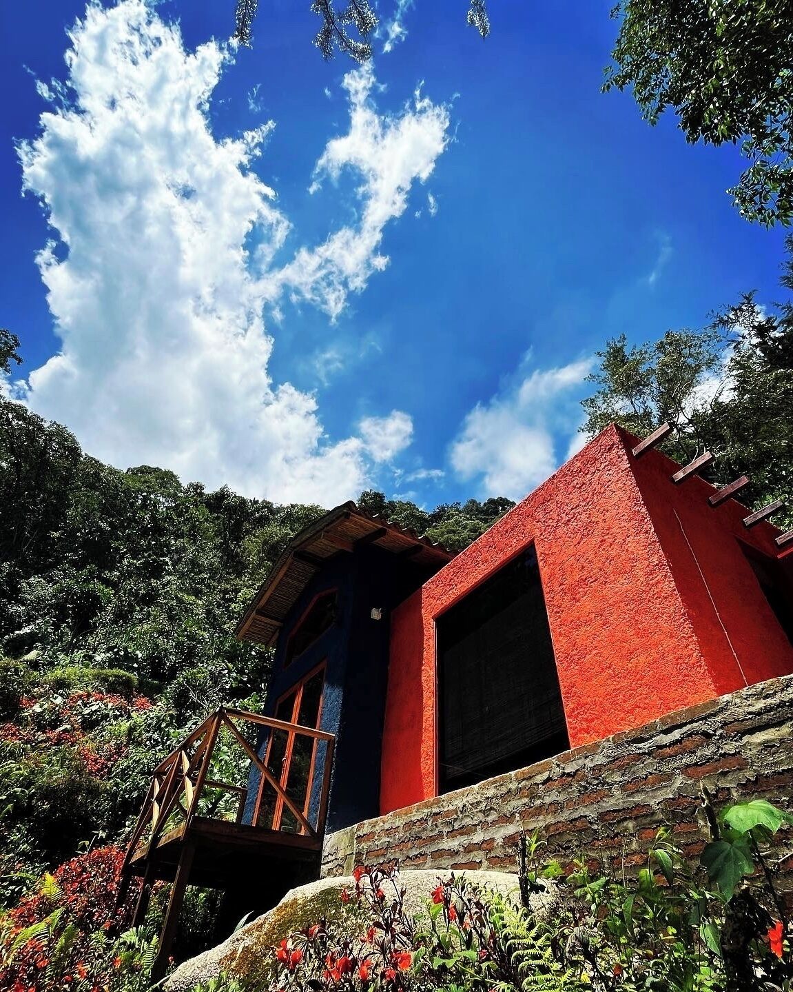 A red-roofed building with a wooden balcony, surrounded by lush greenery and a clear blue sky.