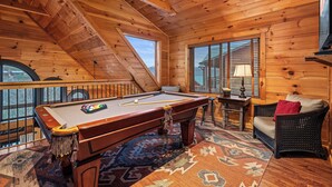 A wooden interior with a pool table, a wicker chair, and a window with blinds. - Beautiful Views, Spacious, Hot Tub, Easy Access Roads, & Free Attractions! (Sevierville)