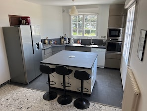A modern kitchen with a refrigerator, oven, and sink. There are four bar stools and a window with a view of greenery.