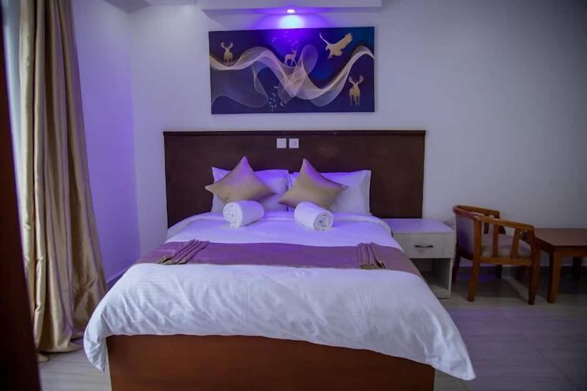 A neatly made bed with a headboard, a wooden chair, a small table, and a painting on the wall.