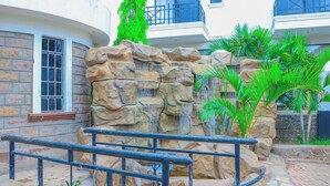 A landscaped area with a rock formation and a waterfall, surrounded by palm trees and a building with balconies. - Asumbi Resort  (Asumbi)