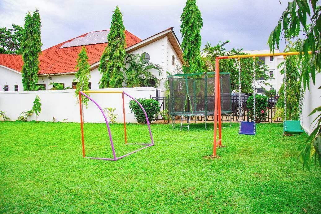 A grassy outdoor area with playground equipment, a white building with a red roof, and tall trees.