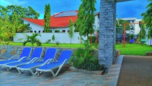 A pool area with blue lounge chairs, a stone wall, and a building with a red roof in the background. - Asumbi Resort  (Asumbi)