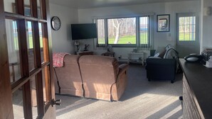 Ein Wohnzimmer mit braunem Ledersofa, einem Fernseher, einer Uhr und einem Fenster mit Blick auf Bäume.