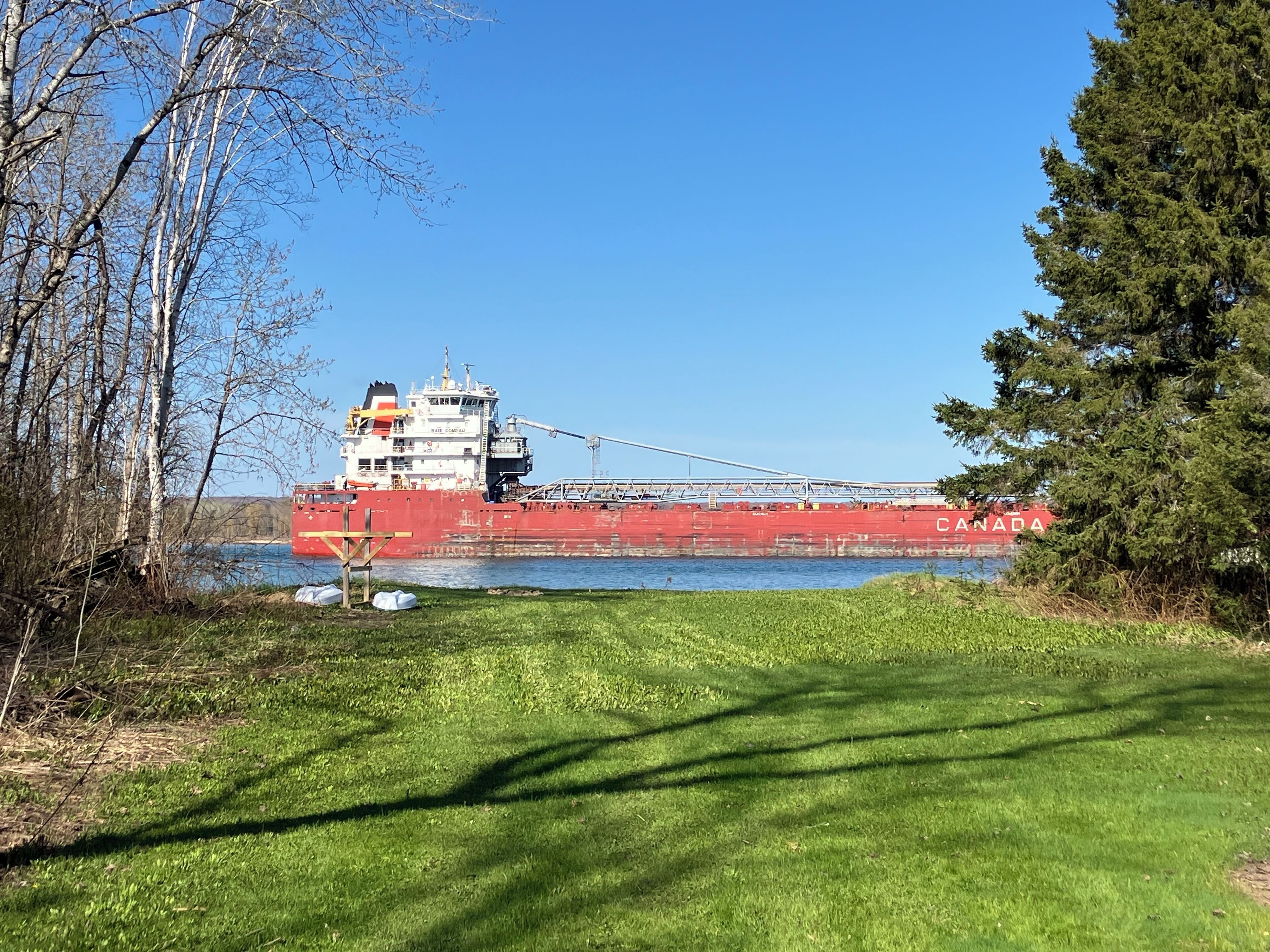 A large red cargo ship with "CANADA" written on its side, docked near a grassy area with trees and a clear blue sky.