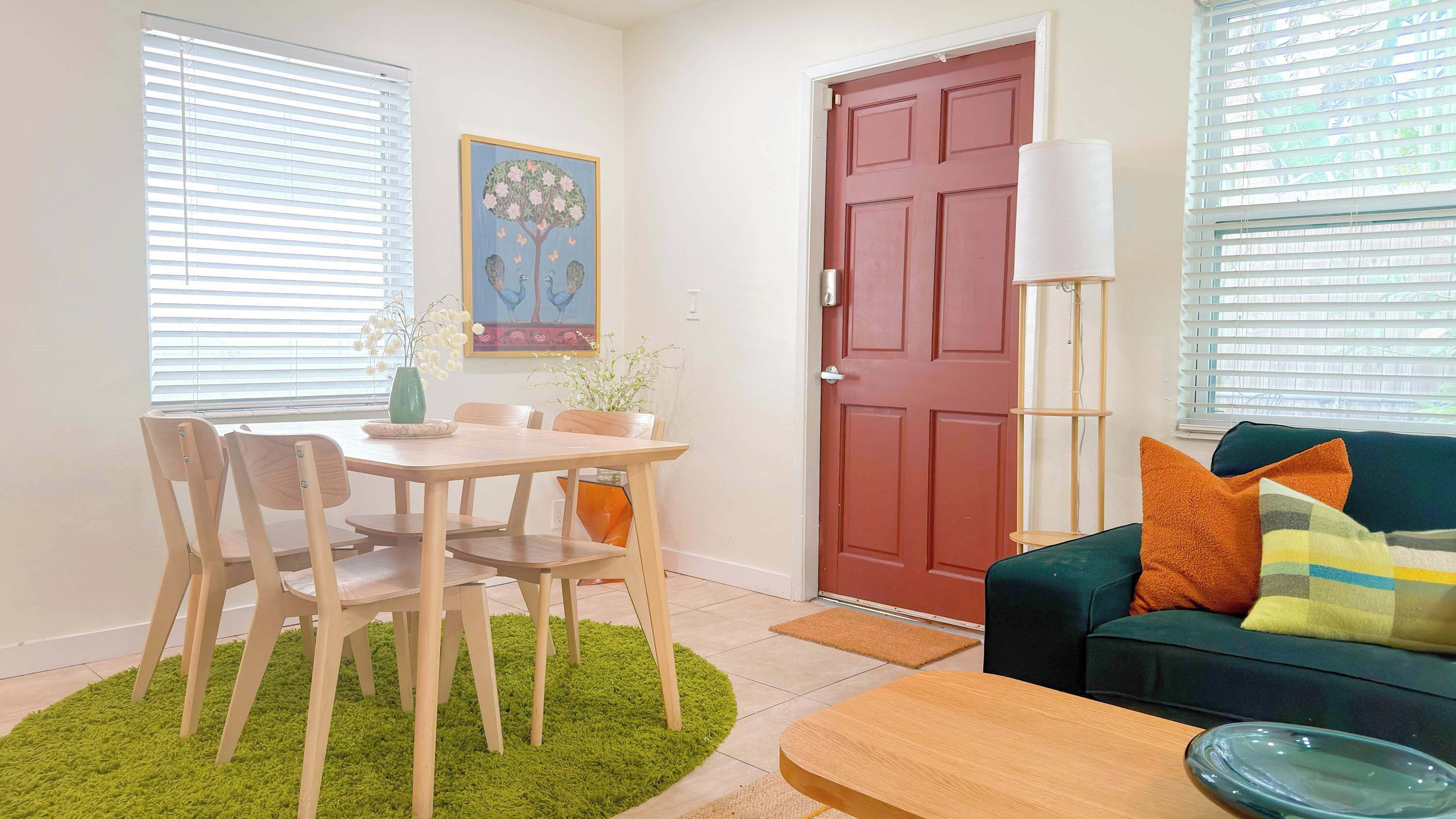 A living room with a dining table, chairs, a sofa, a coffee table, a red door, and a floor lamp.