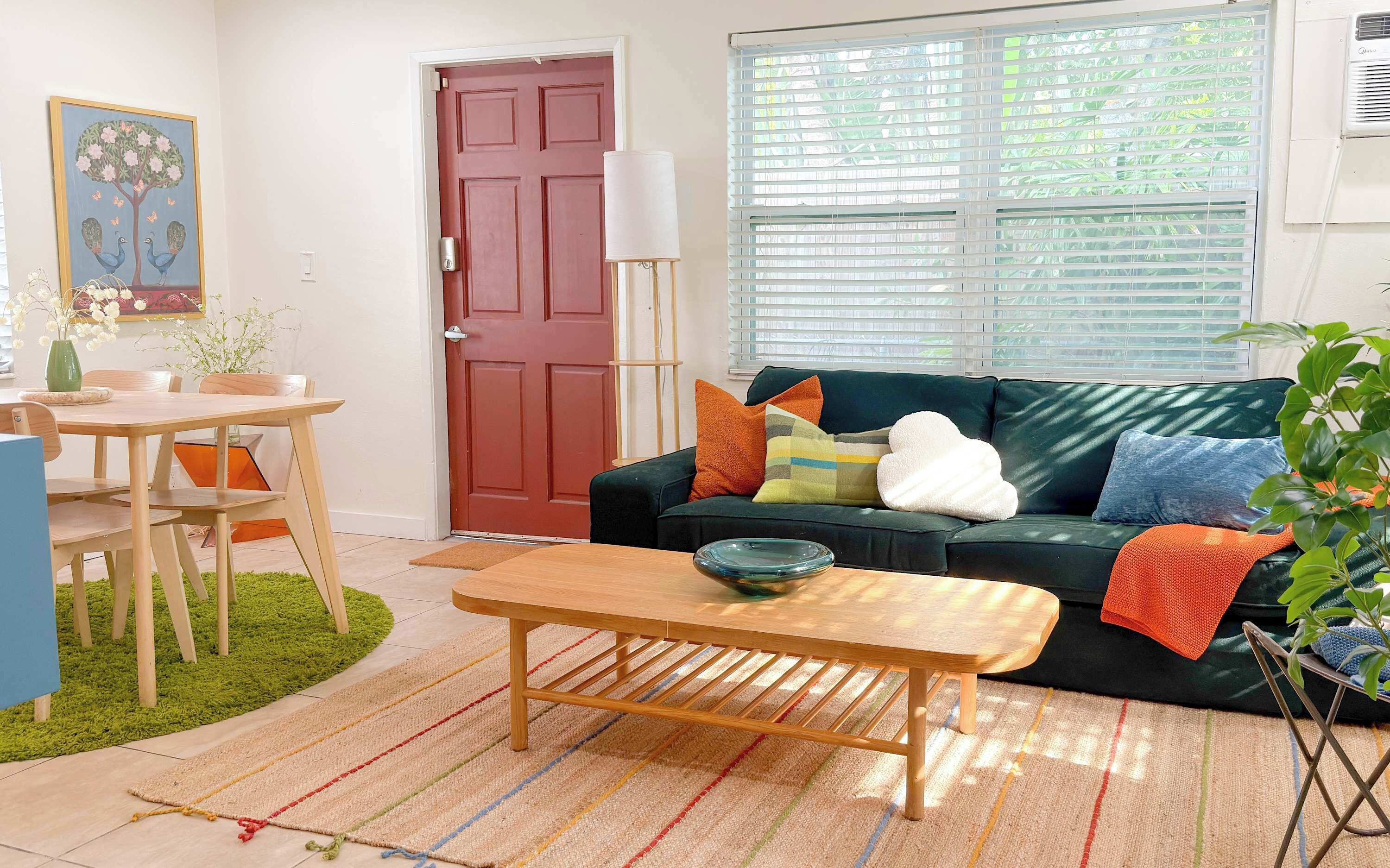 A living room with a dark green sofa, a wooden coffee table, a red door, and a window with blinds.