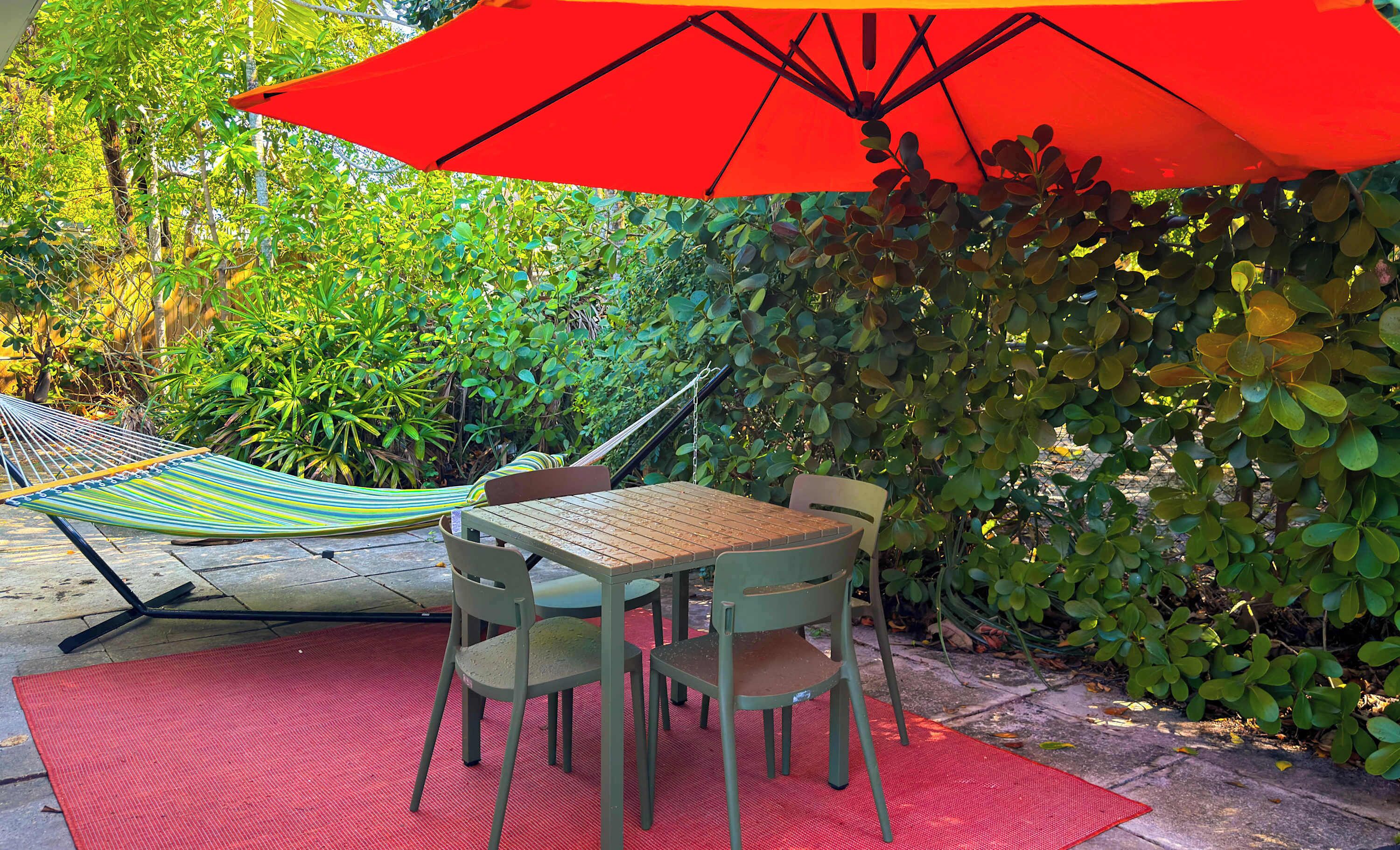 A hammock, a table, two chairs, and a red umbrella in a garden setting.