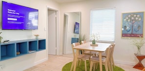 A hotel room with a TV displaying a welcome message, a dining area with a table and chairs, a mirror, and a painting of birds on the wall.