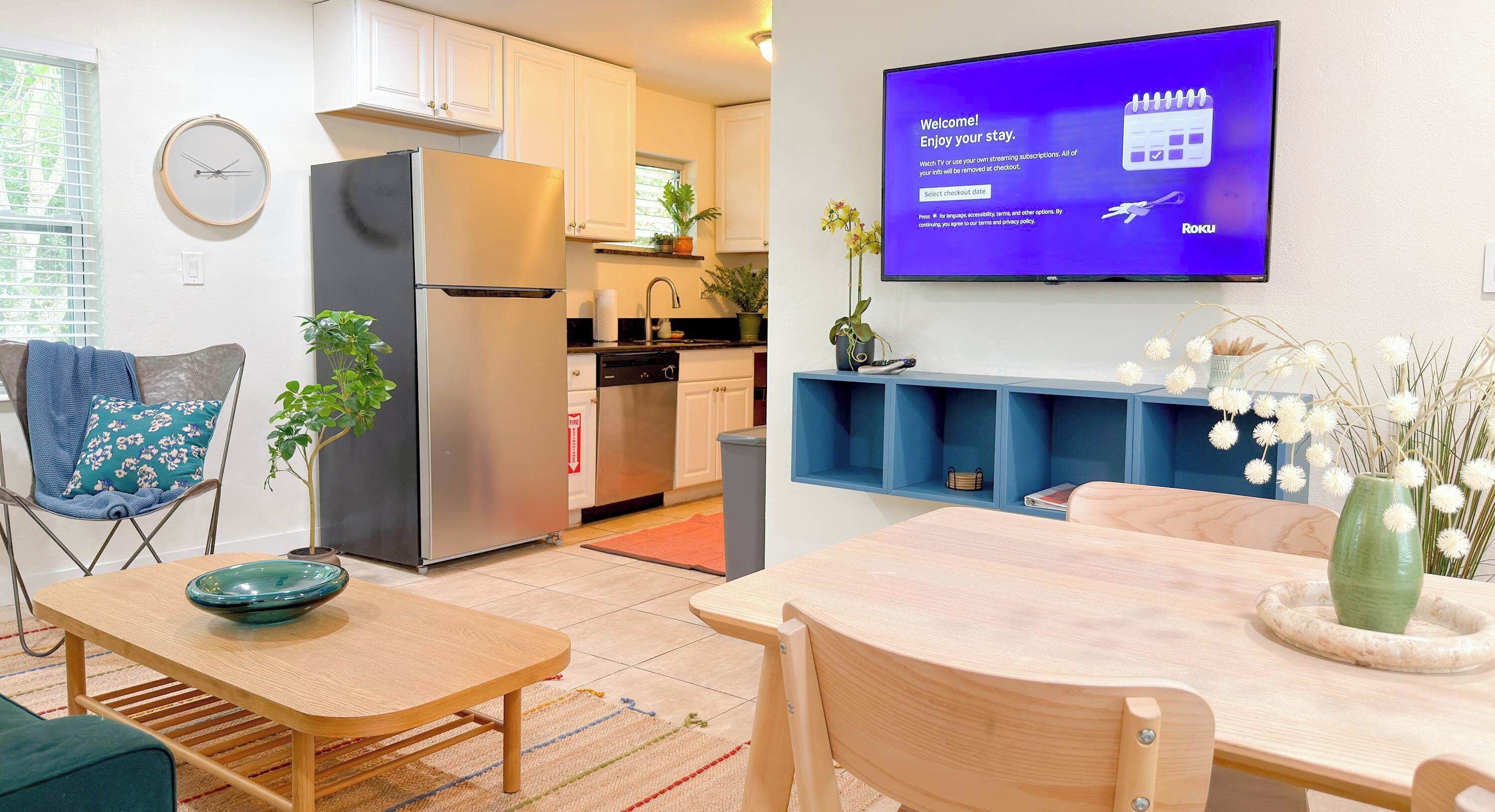 A modern kitchen with a refrigerator, sink, and cabinets, a dining area with a table and chairs, and a TV displaying a welcome message.
