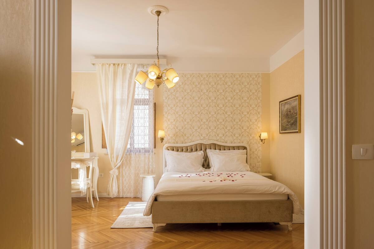A bedroom with a large bed, a chandelier, a vanity with a mirror, and a window with curtains.