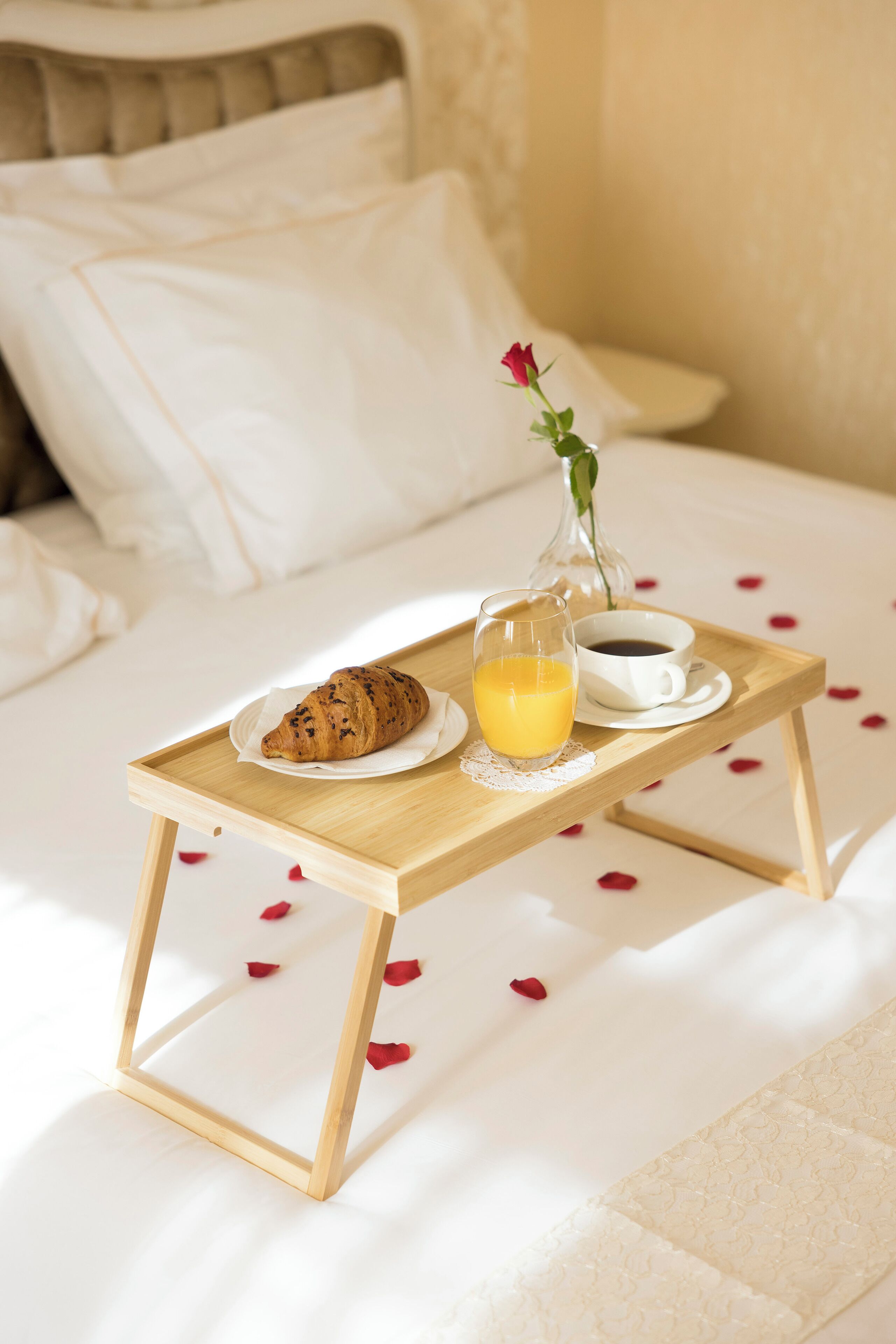 A breakfast tray with croissant, orange juice, and coffee on a bed with white bedding and rose petals scattered around.