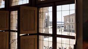Une vue à travers une fenêtre à carreaux, qui révèle une place en plein air avec une statue, un bâtiment et un ciel dégagé.