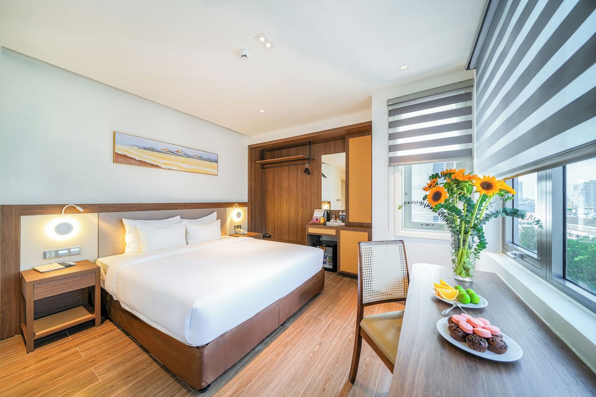 Une chambre d’hôtel avec un grand lit, des tables de chevet, un bureau avec une chaise, un vase de fleurs et une vue sur la ville par la fenêtre.
