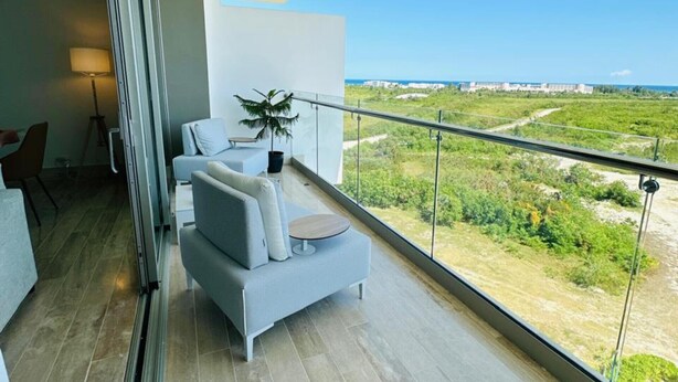 Un balcón moderno con un sofá gris, una mesita pequeña y una planta en maceta. El balcón ofrece vistas a un paisaje verde y un horizonte urbano distante.