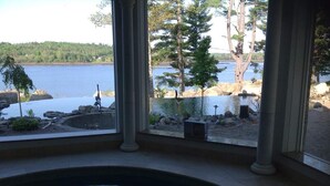 Blick aus einem Zimmer mit großem Fenster auf einen See, ein Steinbecken und eine angelegte Grünfläche mit Bäumen und Felsen.