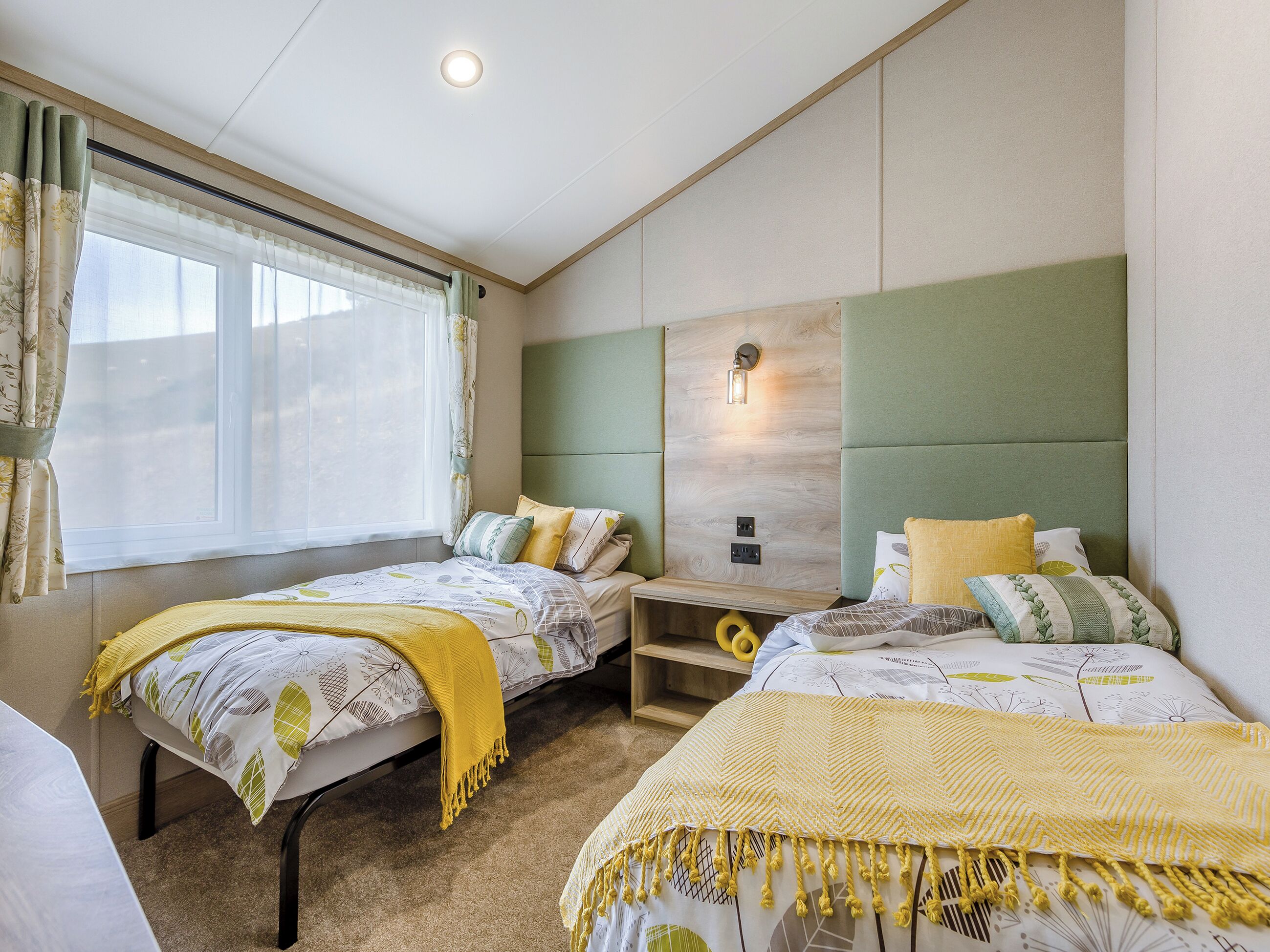 A bedroom with two beds, a window with curtains, a bedside table, and a wall-mounted light.