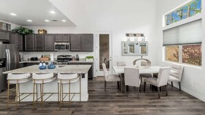 A modern kitchen with dark cabinets, a central island with bar stools, a dining area with a white table and chairs, and a large window with a view.