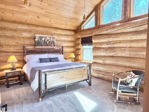 A log cabin bedroom with a wooden bed, two bedside lamps, a rocking chair, and a large window.