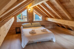 A cozy attic bedroom with a wooden ceiling, a large bed, two windows, and a wall-mounted light fixture.
