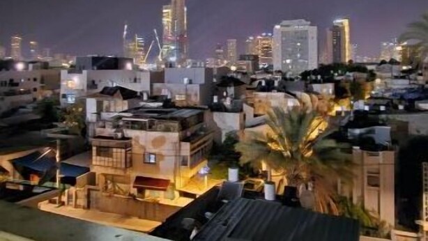 A nighttime cityscape with illuminated buildings and a view of a residential area with houses and a palm tree.