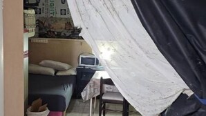 A small, compact room with a bed, a desk, and a chair. There is a window with a curtain partially drawn.