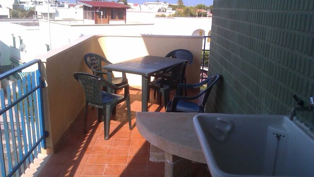 Un balcón con mesa y sillas, un pequeño lavamanos y vistas a edificios al fondo.