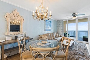 Uma sala de jantar com mesa de vidro e cadeiras de vime, um lustre, um espelho decorativo e vista para o exterior.