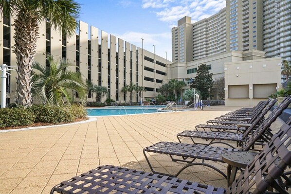 Uma área à beira da piscina com espreguiçadeiras, piscina e um edifício com vários andares e varandas.