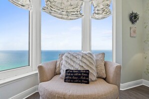 Quarto com vista para o oceano através de amplas janelas, uma poltrona bege com almofada decorativa e uma parede com uma pequena planta e um quadro emoldurado.
