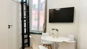 Ein Essbereich mit einem runden Tisch, vier weißen Stühlen, einem an der Wand befestigten Fernseher und einer Schiebetür mit schwarzem Rahmen.