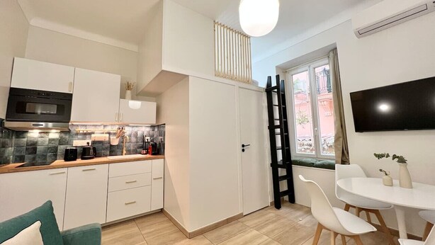 Eine moderne Küche mit weißen Schränken, einer schwarzen Mikrowelle und einer Holzausziehtischplatte. Es gibt einen Essbereich mit einem weißen Tisch und Stühlen sowie einen an der Wand montierten Fernseher.