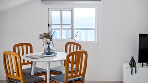 Un coin repas avec une table blanche, quatre chaises en bois, un vase avec des fleurs, une fenêtre donnant sur un paysage agréable et un téléviseur posé sur un meuble blanc.