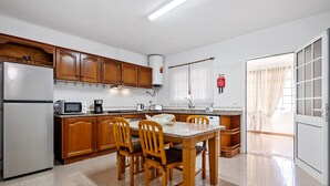 Una cocina con armarios de madera, un refrigerador, un microondas, una mesa con sillas y una ventana con cortinas.