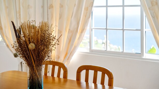 Una mesa de comedor con un jarrón de flores secas, dos sillas de madera y una ventana con vista al mar.