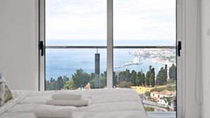 A bedroom with a large window overlooking a coastal cityscape.