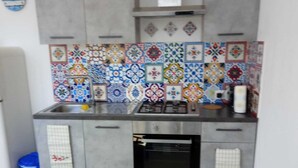 Una cucina compatta con piano cottura a gas, forno e vivace rivestimento in piastrelle. Si nota una sedia blu e un piccolo tavolo con un sottotavolo blu.