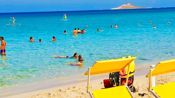 Una spiaggia con acqua cristallina, persone che nuotano e si rilassano al sole, e sedie a sdraio gialle.