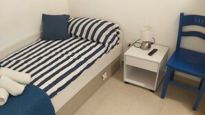 Una piccola camera con un letto singolo, una coperta a righe, un comodino con una lampada e una sedia.