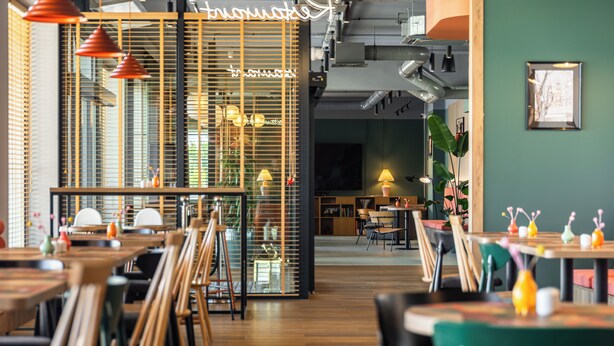 Un restaurant avec des tables et des chaises en bois, une cloison en verre ornée d’un néon, et un mur vert agrémenté d’un tableau encadré.