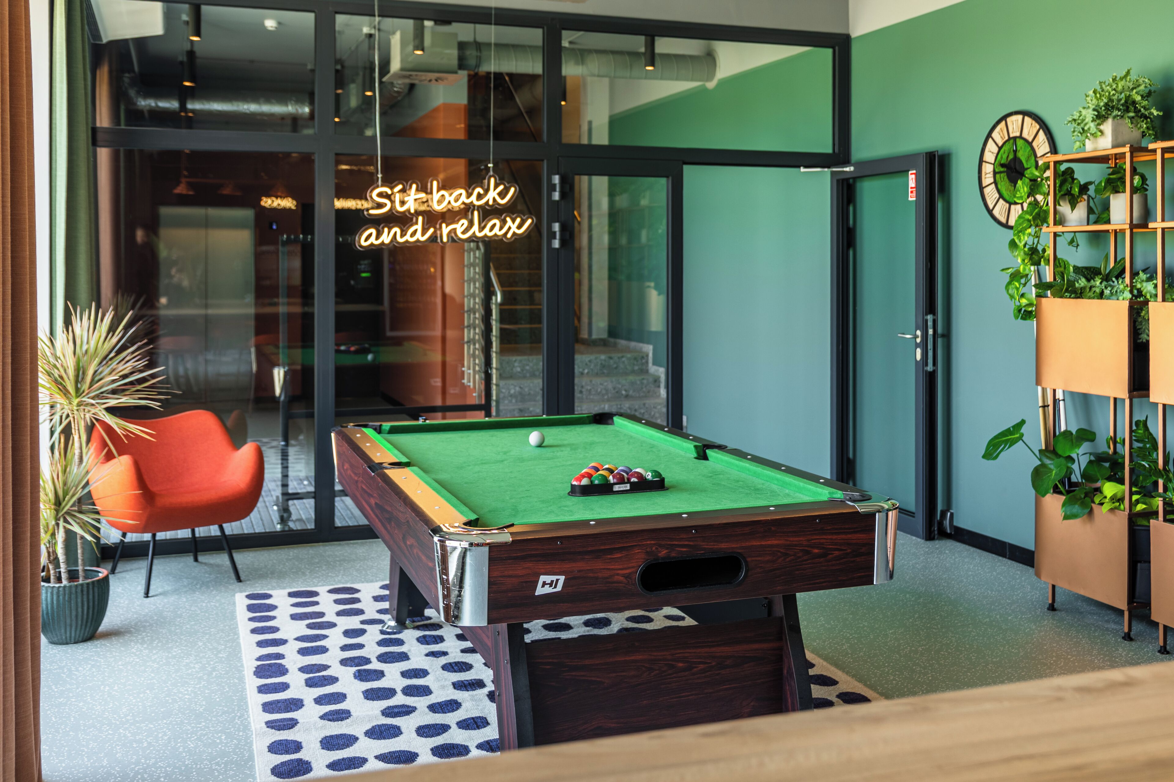 A pool table with a neon sign, a red chair, and a potted plant.